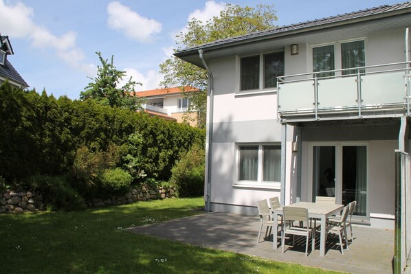 Une maison à deux étages avec un balcon, une terrasse avec une table et des chaises, et un mur de pierre agrémenté de verdure.