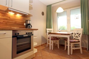 Een kleine keuken met een inbouwoven en koffiezetapparaat op het aanrecht. Er is een eettafel met stoelen en een raam met gordijnen.