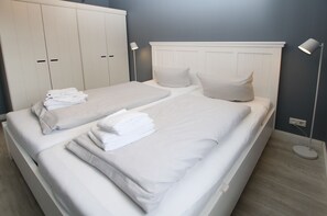 Uma cama de casal com roupa de cama branca, uma mesinha de cabeceira, um guarda-roupa e duas luminárias de chão.
