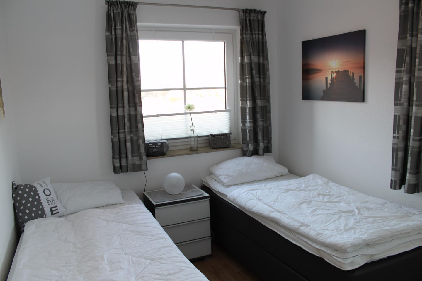 A bedroom with two beds, a nightstand, a window with curtains, a wall-mounted picture, and a bedside table with a lamp.