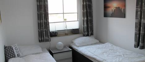 A bedroom with two beds, a nightstand, a window with curtains, a wall-mounted picture, and a bedside table with a lamp.