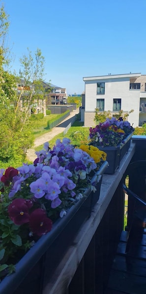 Un balcon orné de jardinières fleuries, d’où l’on peut admirer un bâtiment moderne et la verdure environnante.