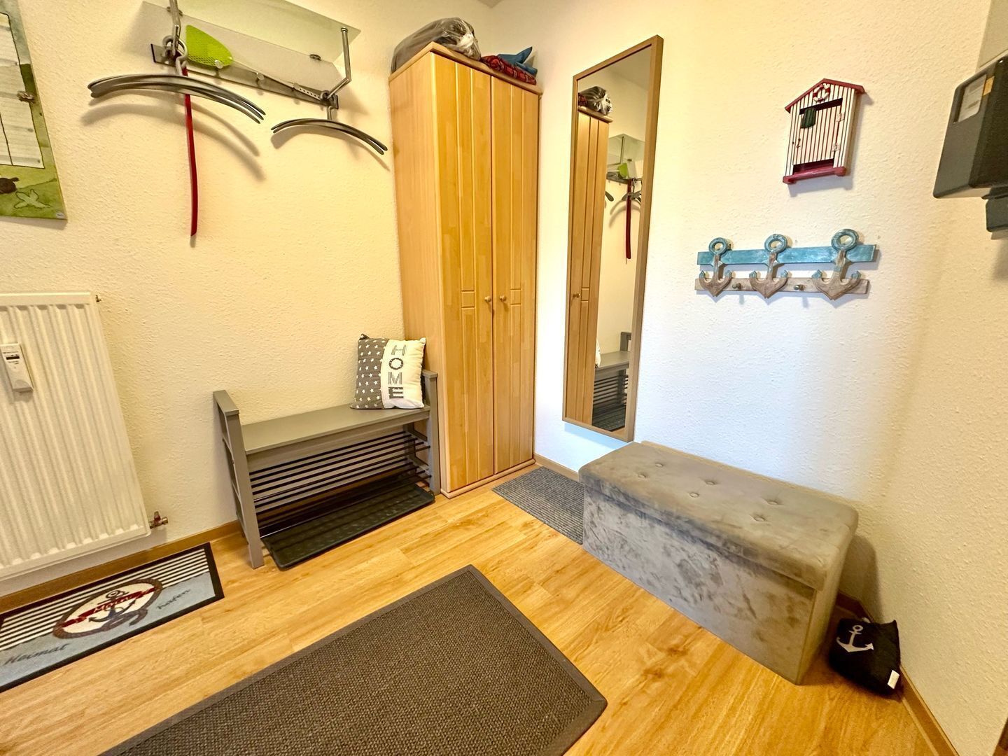 A compact room with a wooden wardrobe, a bench, a small stool, a radiator, a mirror, and wall-mounted hooks.