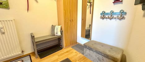 A compact room with a wooden wardrobe, a bench, a small stool, a radiator, a mirror, and wall-mounted hooks.