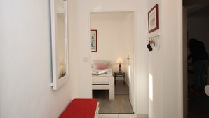 A narrow hallway with a bed in the background, a small table with a lamp, and a coat rack with items hanging. - Sandwall 2, Whg. 4 - Apartment at Sandwall for 2 persons on approx. 44 m² (Wyk auf Föhr)