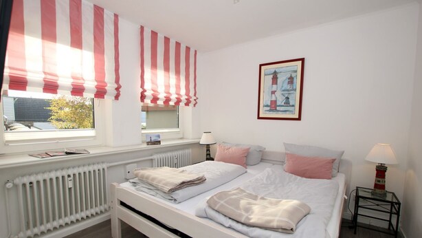 A bedroom with a large bed, two bedside tables, a window with striped curtains, a radiator, and a painting on the wall. - Sandwall 2, Whg. 4 - Apartment at Sandwall for 2 persons on approx. 44 m² (Wyk auf Föhr)