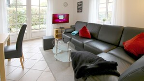 Ein modernes Wohnzimmer mit einer Couch, einem Couchtisch und einem Fernseher. Ein Fenster bietet Blick auf Grünflächen.