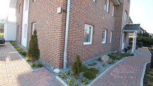 Een gebouw van rode bakstenen met een witte deur en ramen, een verharde oprit en een kleine tuin met struiken en stenen.