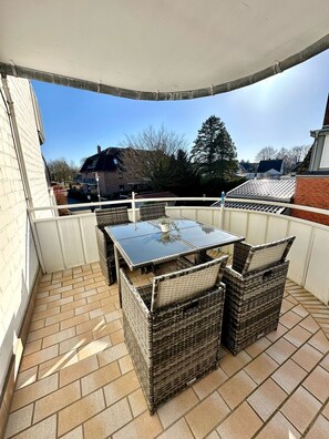 Een overdekt terras met een tafel en rieten stoelen, een betegelde vloer en uitzicht op omliggende huizen.