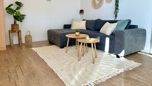 Un soggiorno moderno con un divano grigio, due tavolini in legno, un tappeto bianco e una pianta grande.