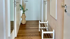 Un corridoio con pavimento in legno, una porta bianca con oblò tondo, una rastrelliera per scarpe a sospensione e una piccola panca bianca.