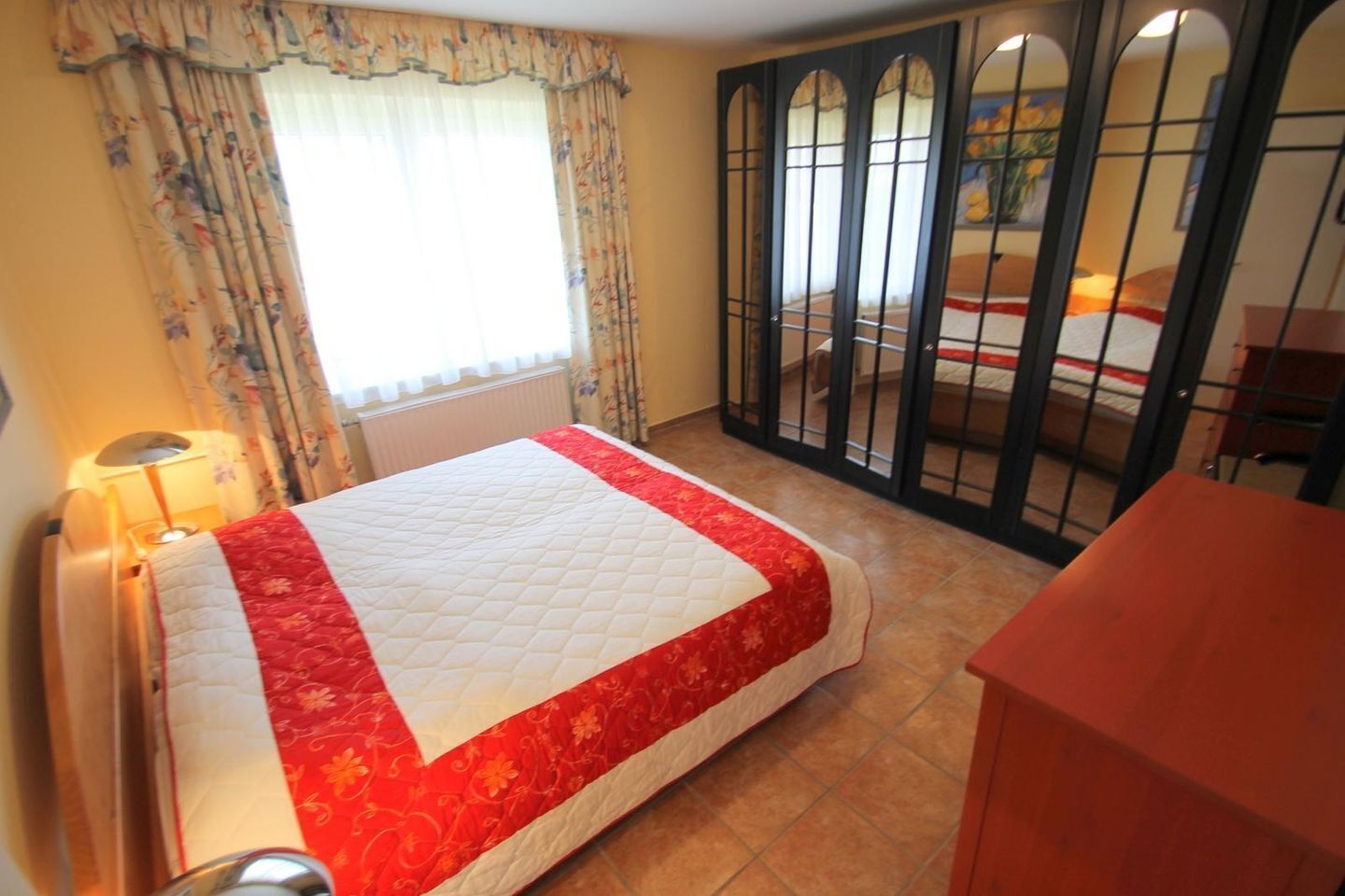 Quarto de hotel com cama, escrivaninha, cadeira, abajur e vista pela janela com cortinas.