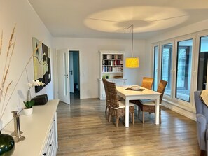 Un espace repas moderne avec une table blanche, des chaises en osier et une bibliothèque en arrière-plan.
