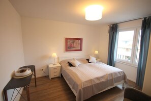 A bedroom with a bed, bedside tables, a desk, a chair, a window with curtains, and a painting on the wall. - 1908 - Am Kurpark - 1908 - Apartment on the 2nd floor "Am Kurpark" (Dahme)