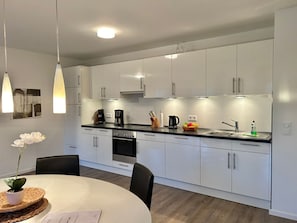 Eine moderne Küche mit weißen Schränken, einer schwarzen Arbeitsplatte und Edelstahlgeräten. Es gibt einen Esstisch mit Stühlen und einen Blumentopf.
