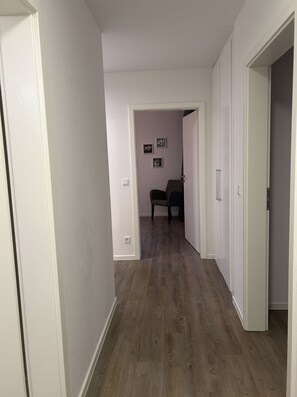 Un pasillo con suelo de madera, paredes blancas y una puerta que da a una habitación con una silla.