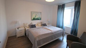 A bedroom with a bed, bedside tables, a lamp, a painting, a window with curtains, and a chair.