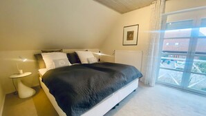 A bedroom with a large bed, a bedside table with a lamp, a radiator, and a window with a view of buildings.