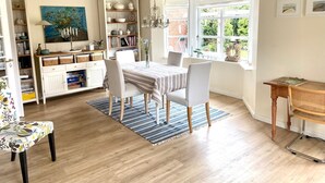 A bright, open-plan living space with a dining area, bookshelves, and a view of greenery through large windows.