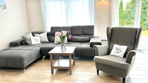 A modern living room with a grey sectional sofa, a grey armchair, and a small table with a vase of flowers. - 1310 - House Greta - House Greta in Dahme 1310 (Dahme)