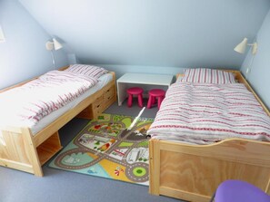 Un dormitorio con dos literas de madera, un escritorio, dos taburetes rosa y una alfombra de juego colorida.