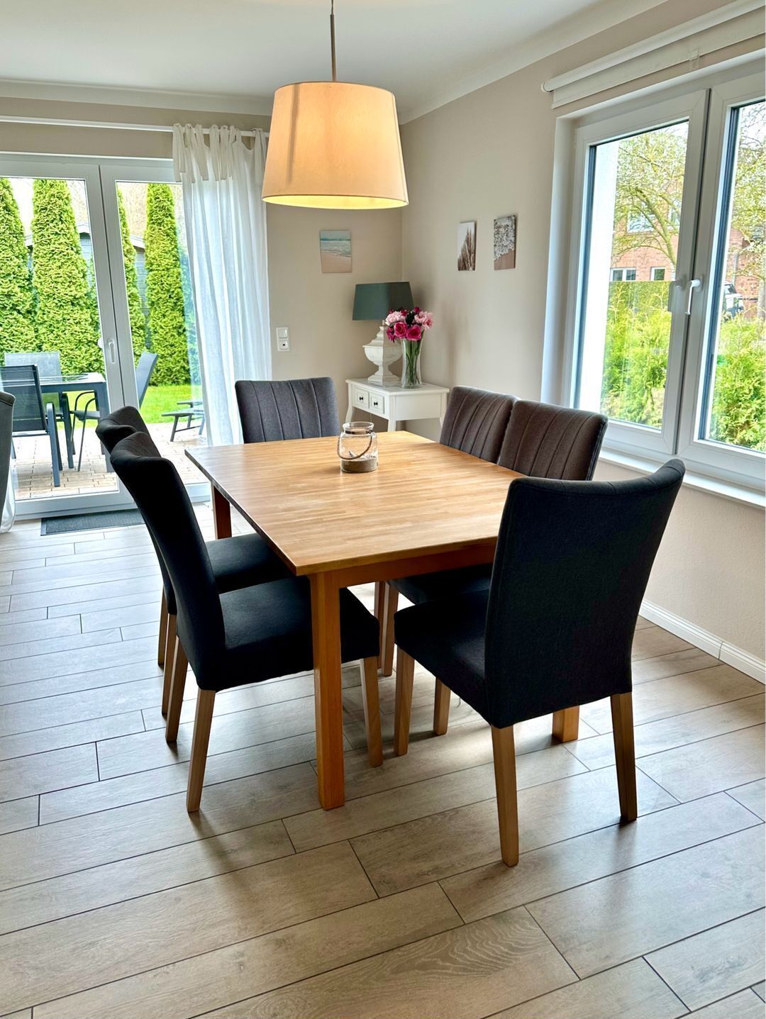 A dining area with a wooden table and black chairs, a hanging lamp, and a view of greenery outside.