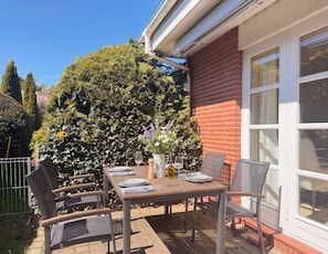 A patio table set for two with chairs, a potted plant, and a window with a view of a brick wall and greenery.
