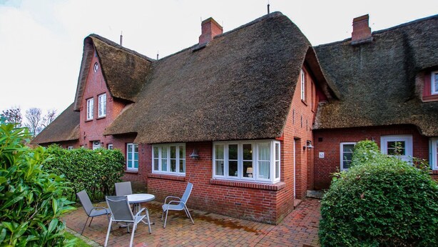 Une maison traditionnelle en briques avec un toit de chaume, un petit patio avec une table et des chaises, et un jardin bien entretenu.