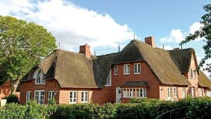 Une maison traditionnelle à toit de chaume, dotée d’une façade en briques, de nombreuses fenêtres et d’une allée gravillonnée.