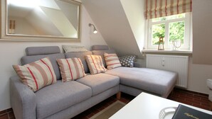 A grey sectional sofa with striped pillows, a white table, a book, a radiator, and a window with a striped valance. - Am Grünstreifen 31, Whg. 6 "Strandgut" - Apartment for 2 persons on approx. (Wyk auf Föhr)