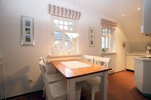 Un comedor con una mesa de madera y sillas blancas, una ventana con cortinas, y una zona de cocina con electrodomésticos blancos.
