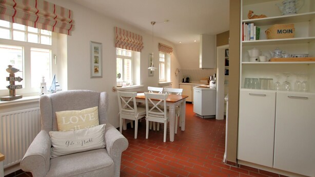 A cozy living room with a grey armchair, a dining table with white chairs, a kitchen area with white cabinets, and a red brick floor. - Am Grünstreifen 31, Whg. 6 "Strandgut" - Apartment for 2 persons on approx. (Wyk auf Föhr)