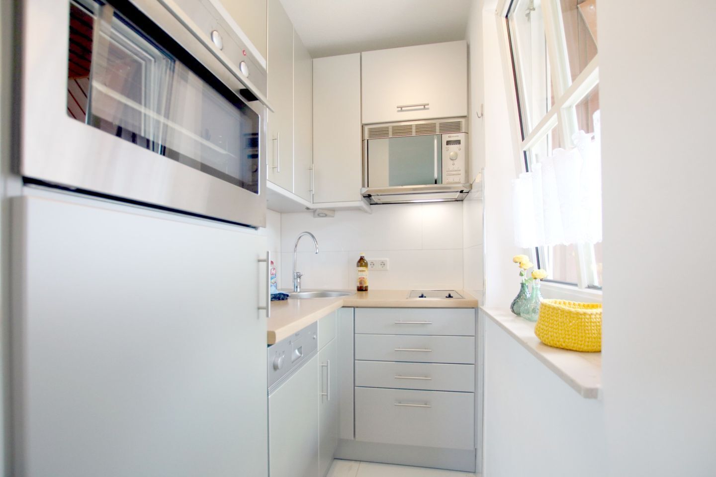 A compact kitchen with a built-in oven, microwave, and sink. There are cabinets and a countertop with a yellow basket on the window sill.