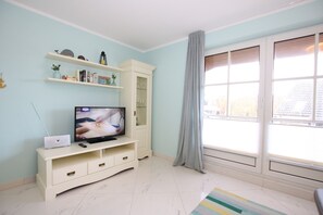 A living room with a flat-screen TV on a white entertainment unit, a bookshelf, a window with blinds, and a striped rug.