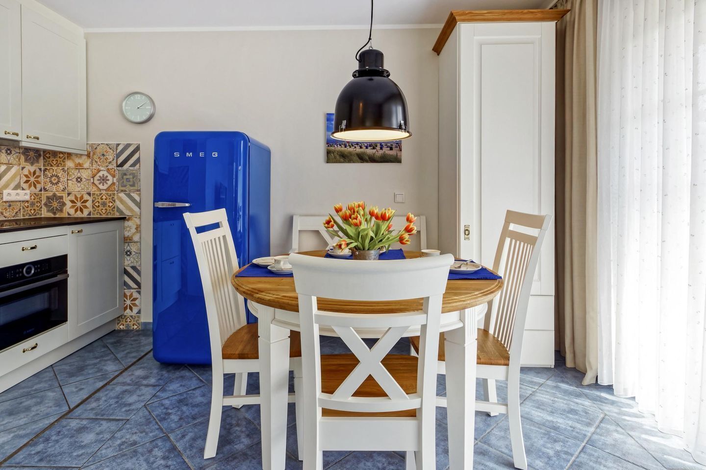 A kitchen with a blue refrigerator, white cabinets, a wooden dining table, and white chairs.