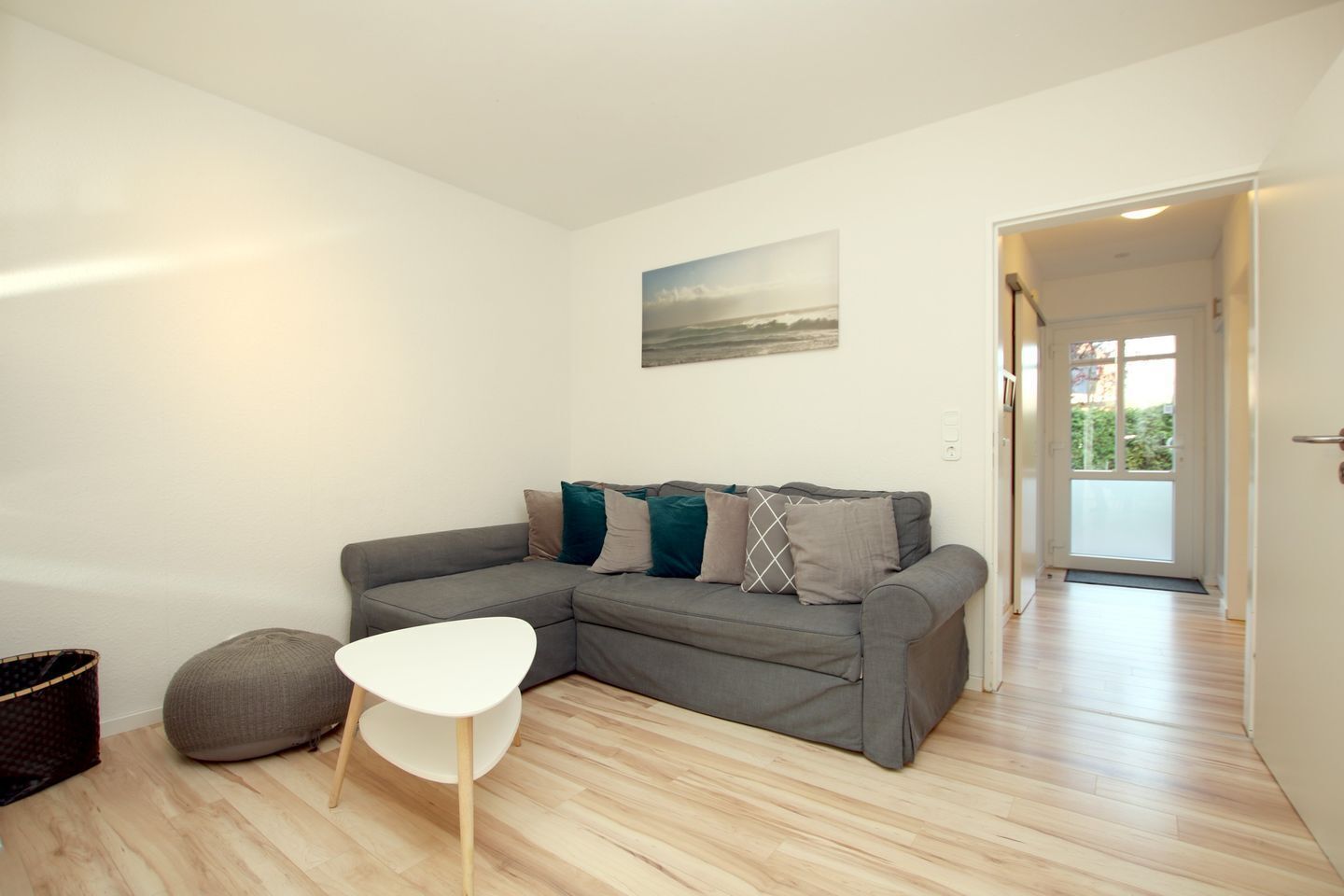 Un salon moderne avec un canapé d’angle gris, une table basse blanche et un pouf gris. Un tableau est accroché au mur, et une porte donne sur un balcon.