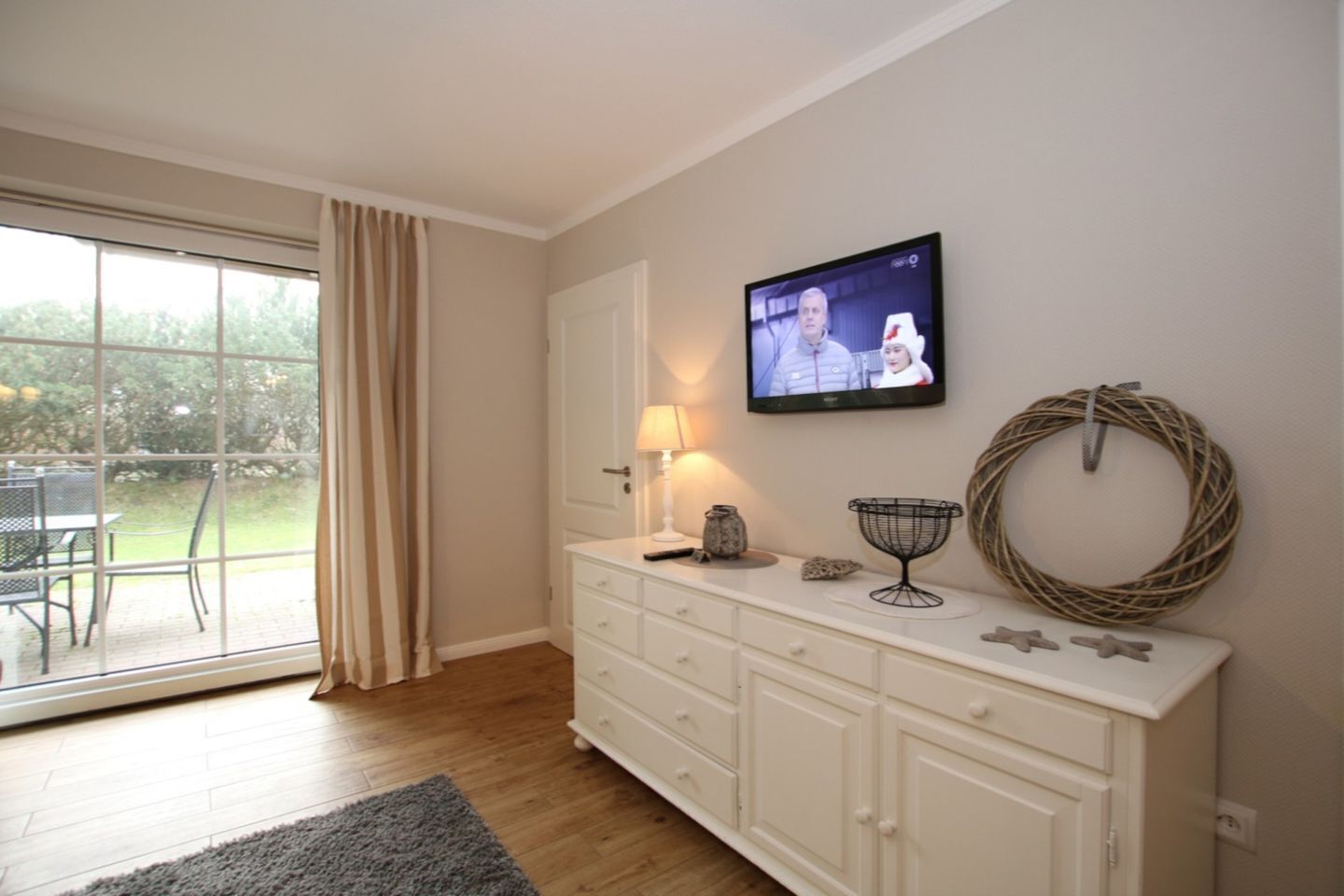 A room with a sliding glass door leading to an outdoor area, a white cabinet with a television mounted above it, and a wicker wreath on the wall.