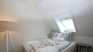 Een slaapkamer met schuine wanden, een bed met gestreept beddengoed, een nachtkastje, een lamp en een raam.