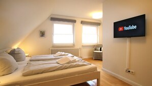 A bedroom with a bed, two windows, a radiator, a chair, and a TV mounted on the wall. - Linge 4, Whg. 1, Hygge - vacation apartment with garden, total of 6 rooms, 4 bedrooms (Wrixum)