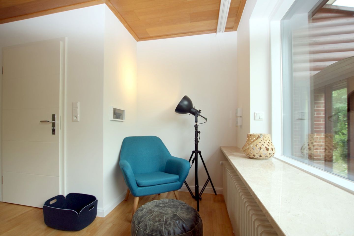 Een kamer met een blauwe fauteuil, een vloerlamp, een mand en een raam met uitzicht op groen.