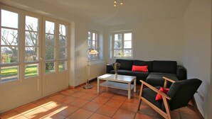 Een ruime woonkamer met een zwarte bank, een witte salontafel en een houten stoel. Grote ramen laten natuurlijk licht de kamer binnen.