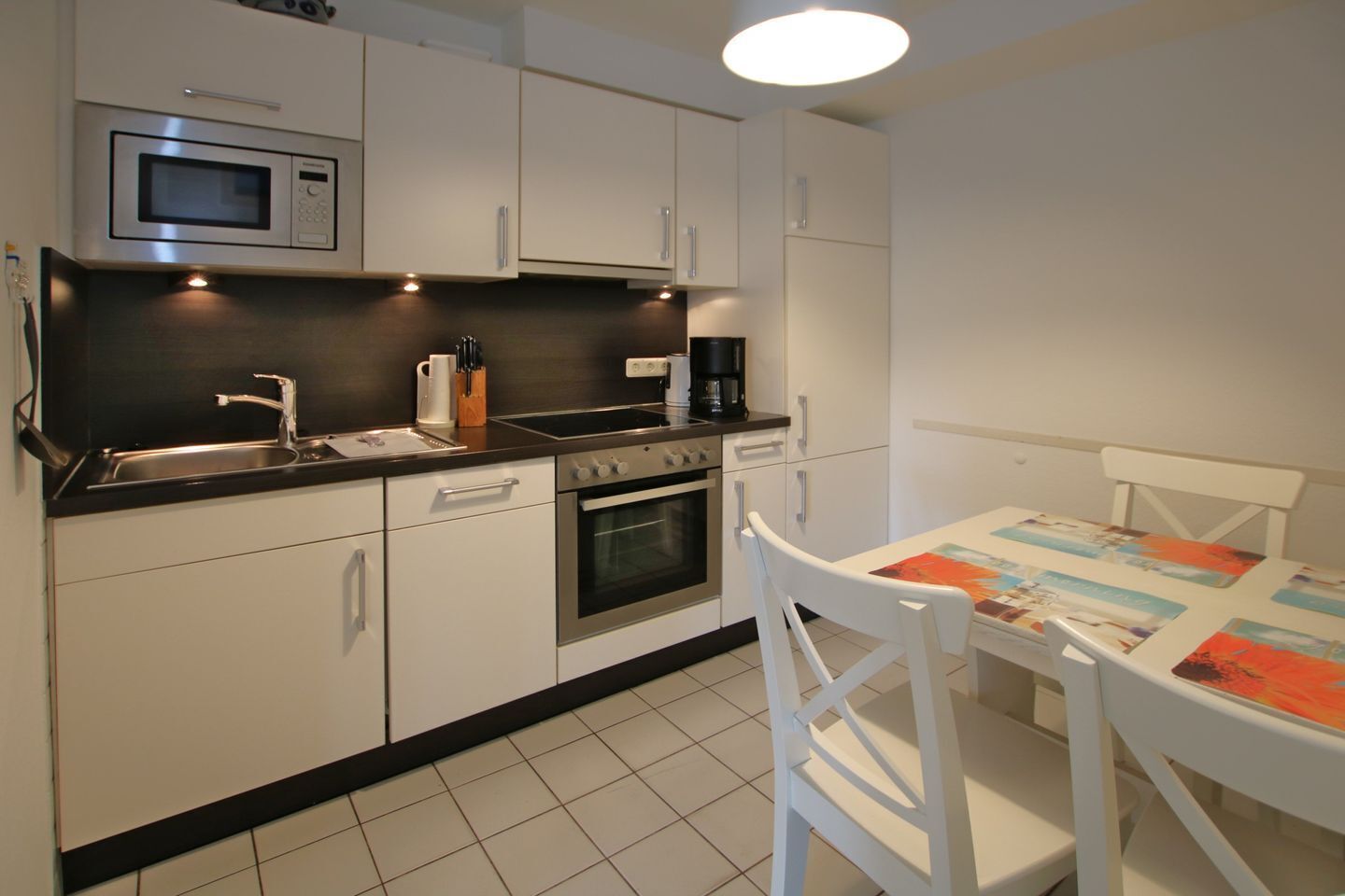Une cuisine compacte avec des meubles blancs, un four à micro-ondes, une cuisinière et une machine à café. On y trouve une petite table à manger avec une nappe colorée et des chaises blanches.