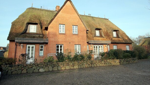 Een twee verdiepingen tellend huis met een flauw hellend dak, veel ramen en een stenen muur ervoor.