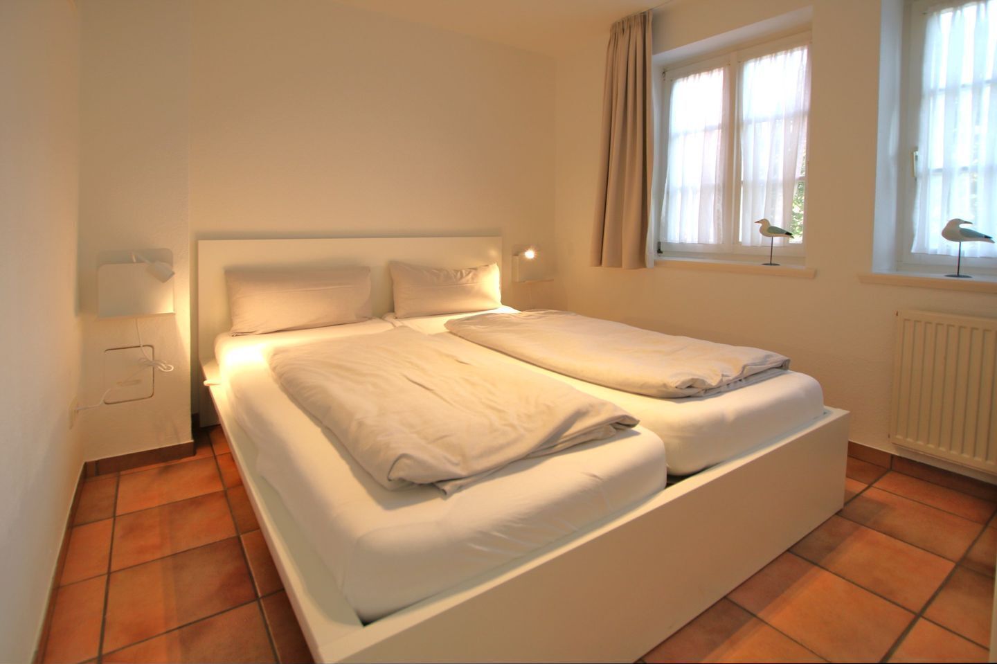 Een tweepersoonsbed met wit beddengoed in een kamer met betegelde vloer en een raam met gordijnen.