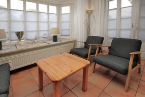 Una habitación con una mesa de madera, dos sillones grises, un radiador y una ventana con persianas.