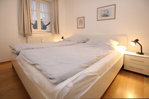 Una cama doble con ropa de cama blanca y almohadas, una mesita de noche con lámpara, y dos cuadros en la pared.