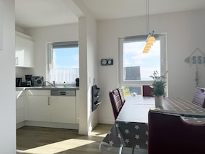 Une cuisine moderne avec des meubles blancs, un coin repas comprenant une table et des chaises, un luminaire suspendu et une fenêtre donnant sur la ville.