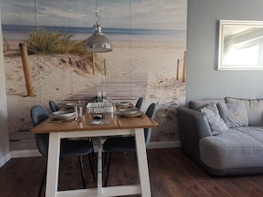 Uma sala de jantar com vista para a praia, uma mesa de madeira posta com pratos e copos, e um sofá cinza na sala de estar.