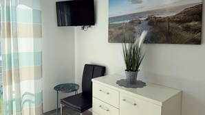A room with a TV mounted on the wall, a painting of a coastal landscape, a chair, a small table, and a cabinet with drawers. - 2605 - Strandwiese 18 - Apartment 2605 in the Strandwiese (Dahme)