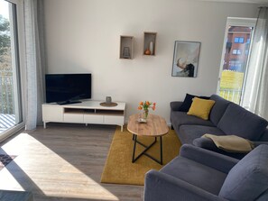 Un salon moderne avec un téléviseur à écran plat posé sur un meuble blanc, un canapé gris agrémenté d’un coussin jaune, une table basse en bois sur laquelle se trouve un vase de fleurs, et un tableau abstrait encadré accroché au mur.